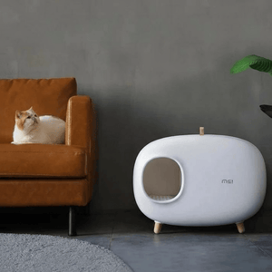 White pet house next to a brown sofa with a cat on it against a gray wall.
